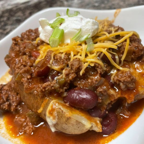 Hearty chili-topped baked potato with shredded cheese, sour cream, and green onions in a white bowl.